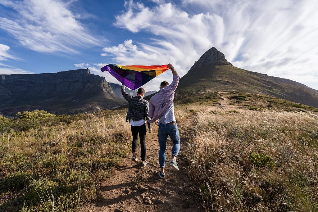 Two people walking on a trail towards Signal Hill & Table Mountain holding the LGBTQIA+ flag