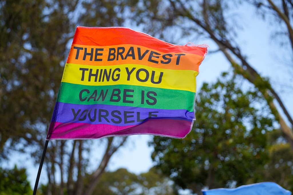 A Pride flag with the text The Bravest Thing You Can Be is Yourself flies in the air during a march